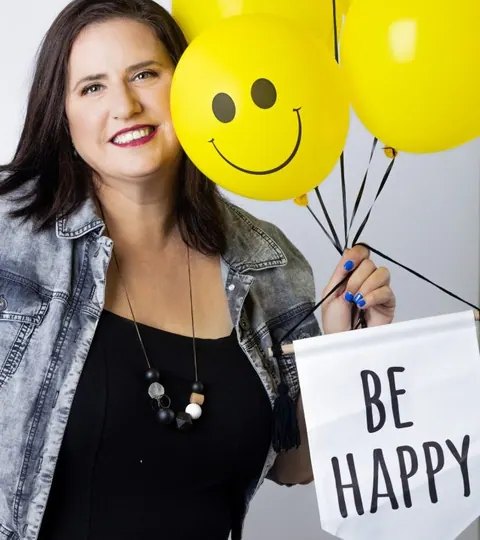 בילי הראל מחזיקה בלונים צהובים ושלט Be Happy